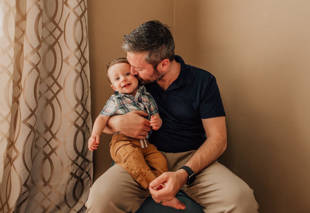 Family photoshoot with dad holding son in lap and giving him a kiss on the cheek.