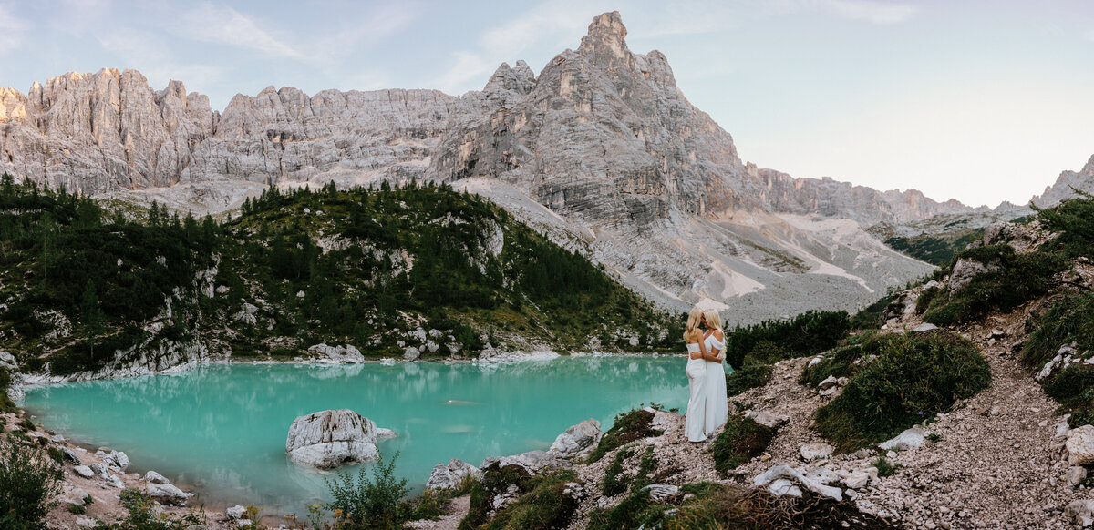 italy dolomites elopement photographer couple stands beside blue lake with mountain background