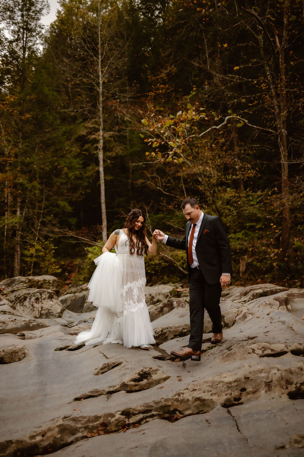 Groom helping the bride carefully walk across smooth rock formations at Greenbrier during their eloping to Gatlinburg, surrounded by dense forest and early autumn colors.
