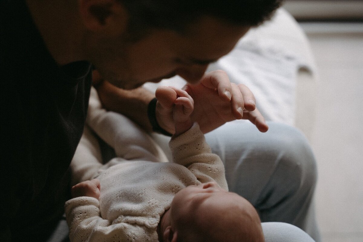 Le père tient la petite main de son bébé endormi et l’embrasse délicatement. Séance photo naissance par Kamescop, vidéaste et photographe en Bretagne.