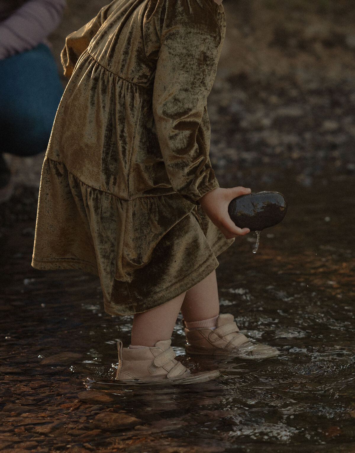toddler wearing a green velvet dress picks up and holds rocks at the river in Bend, Oregon