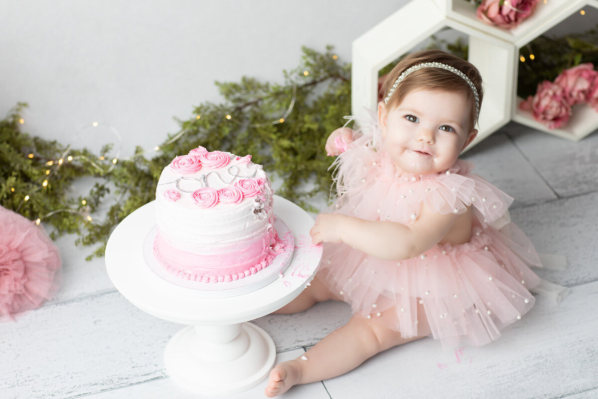 Dallas milestone photographer captures a baby girl in a pearl-trimmed pink tutu dress enjoying a cake smash with pink florals, greenery, and a plush flamingo in a soft, elegant setup.