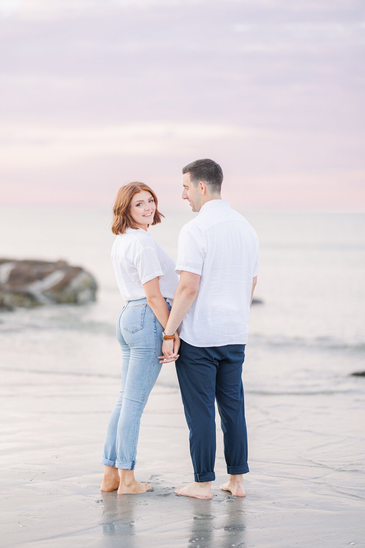 A man and woman holding hands with the woman looking at the camera during their Wingaersheek Beach sunset engagement photos.