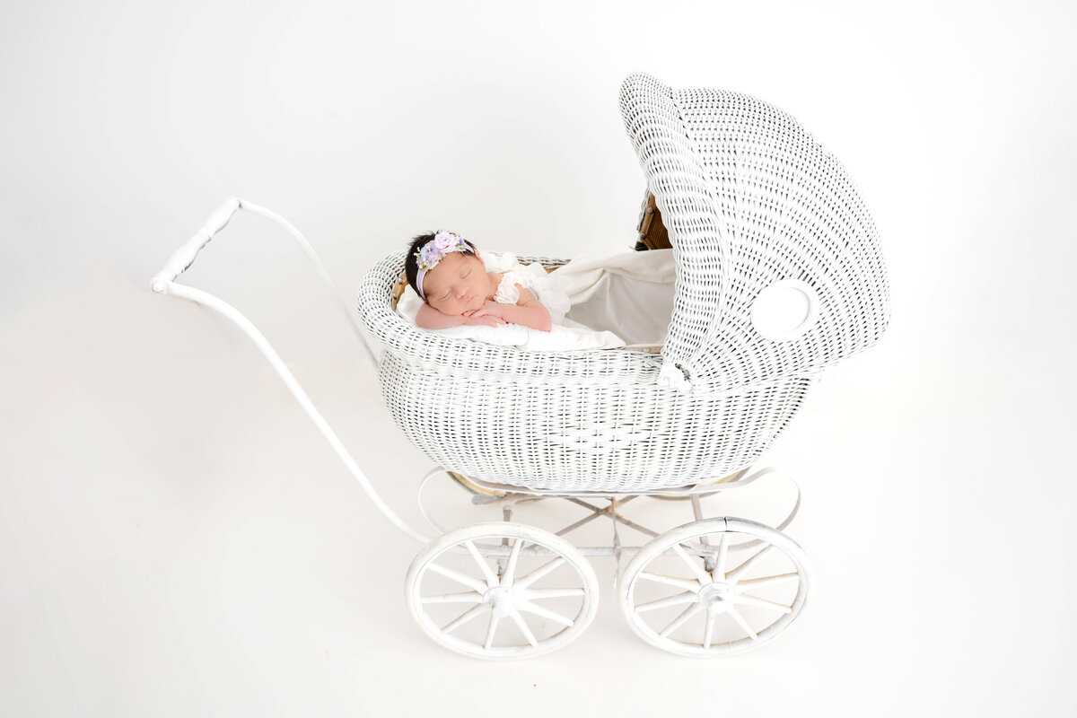 Newborn baby dressed in a flowy white dress inside of a white vintage stroller resting with chin on arms.