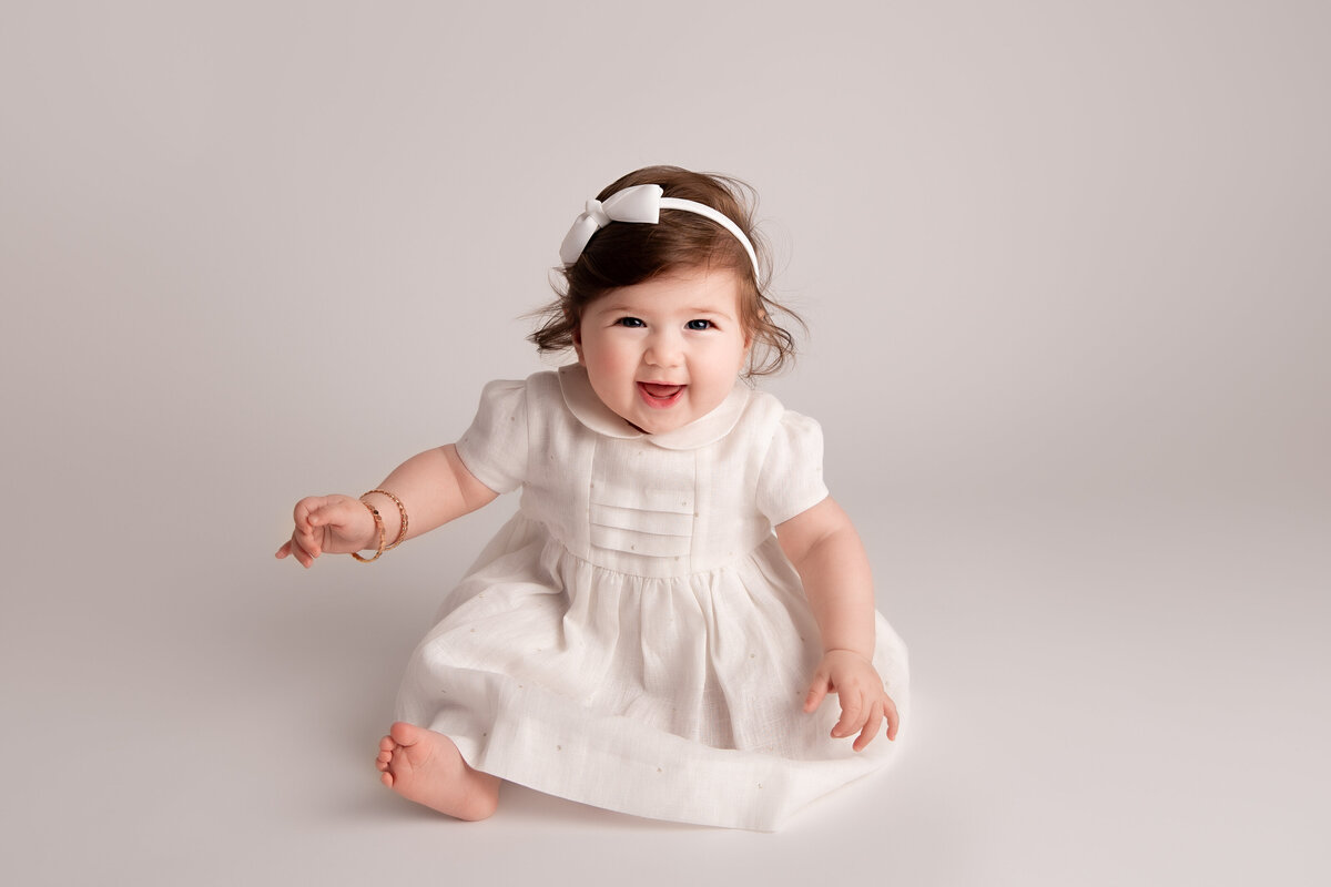 Happy six month old baby girl sitting on a white backdrop wearing a white dress and a bow headband.