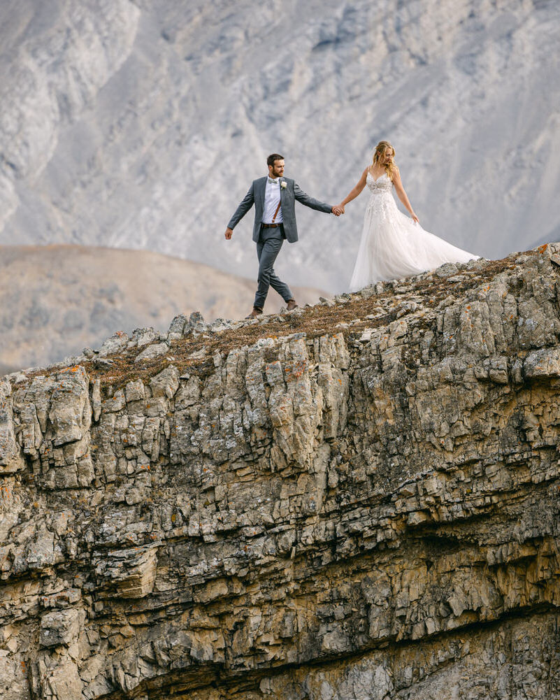 This intimate photograph captures the groom holding the bride's hand as they walk down a slope, having just arrived by helicopter for their private wedding in Banff National Park. The breathtaking landscape adds a sense of adventure and exclusivity to their unforgettable moment.