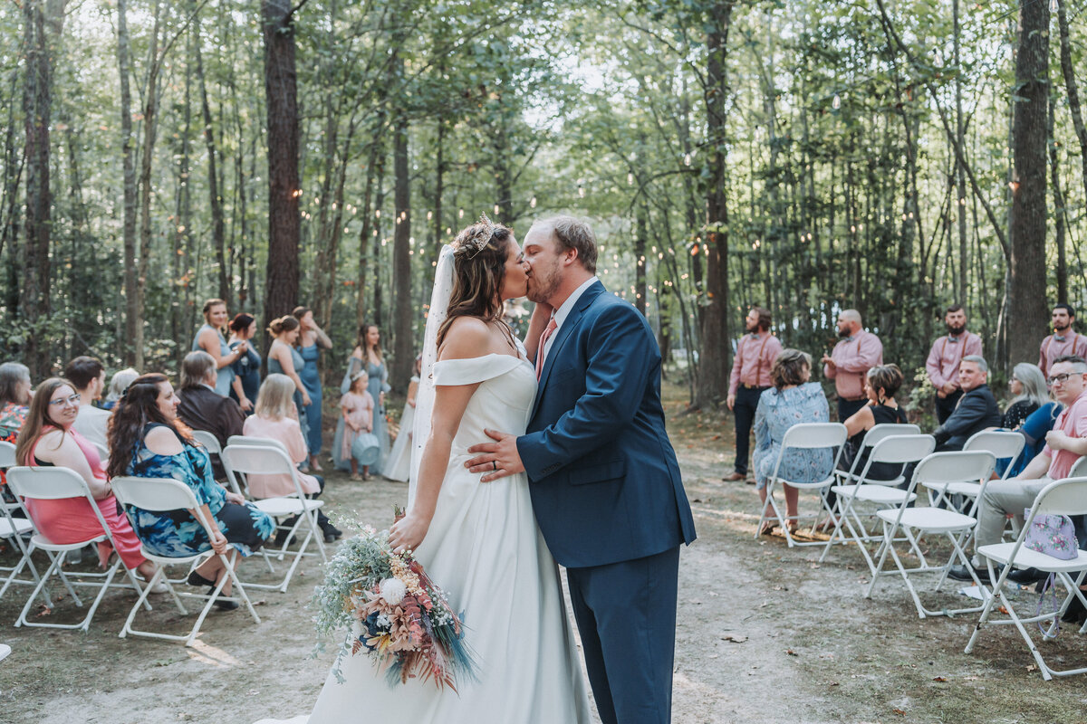 Newlywed couple sharing a kiss during their woodland wedding ceremony, surrounded by guests seated on white folding chairs. The bride holds a bouquet of colorful flowers, and the groom wears a navy suit. Shot by Quinn Photography LLC.
