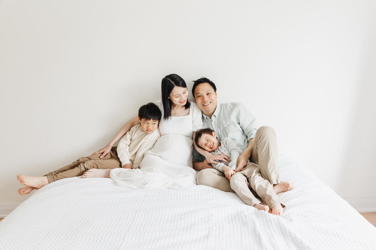 Pregnant mom surrounded by husband and two older sons. They are sitting on a white bed.