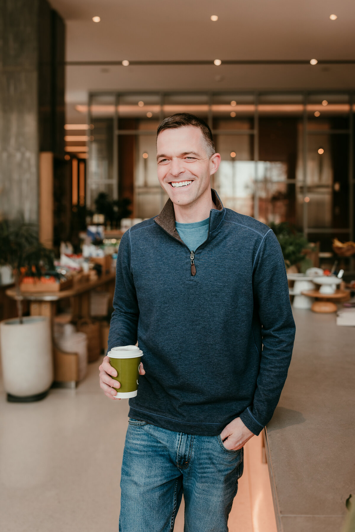 Man in a blue sweater holding a coffee cup in a modern coffee shop, showcasing a casual, confident online dating profile look.