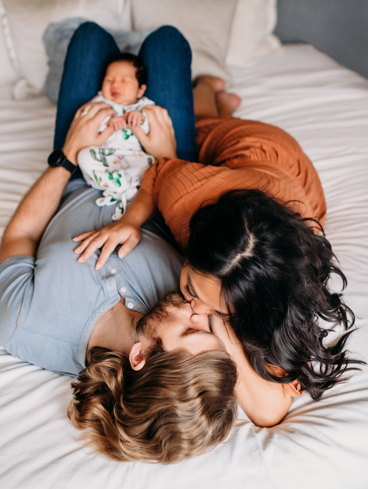 Newborn Photographer, Mom and dad lying down on bed and kissing each other while baby girl stays on dad's lap.