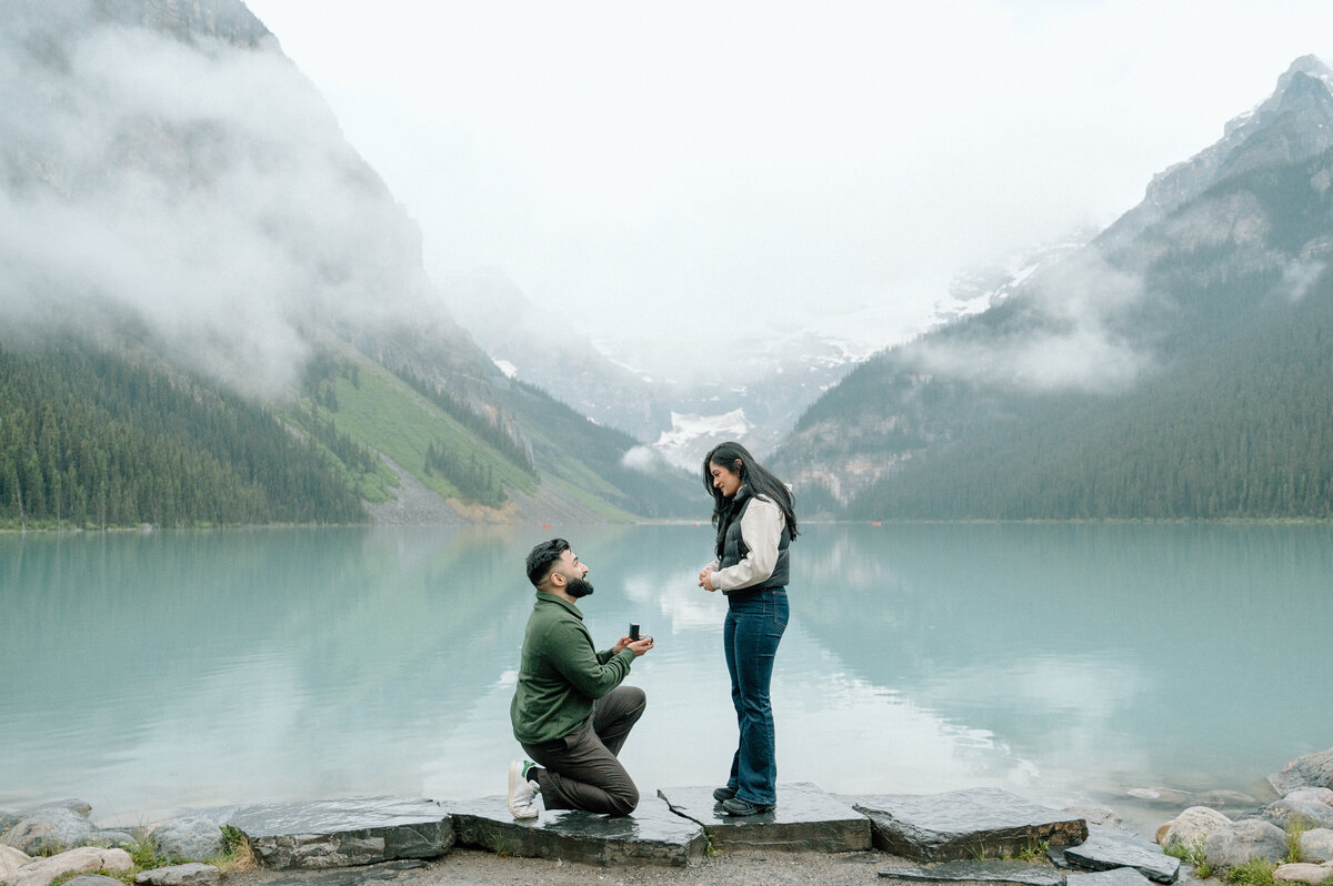 Romantic proposal at Lake Louise with groom-to-be kneeling in front of turquoise lake and mountains.