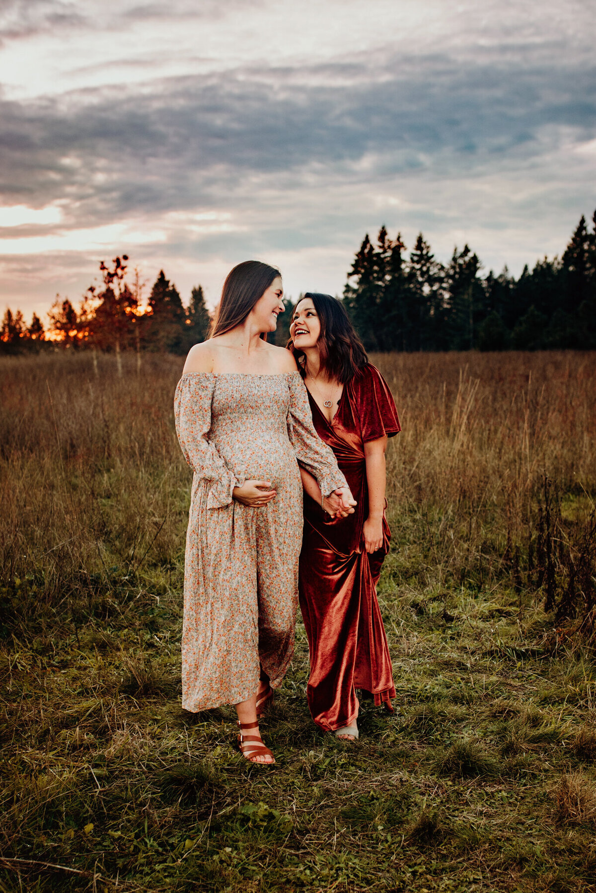 Two expectant moms walking hand in hand through a grassy Oregon field