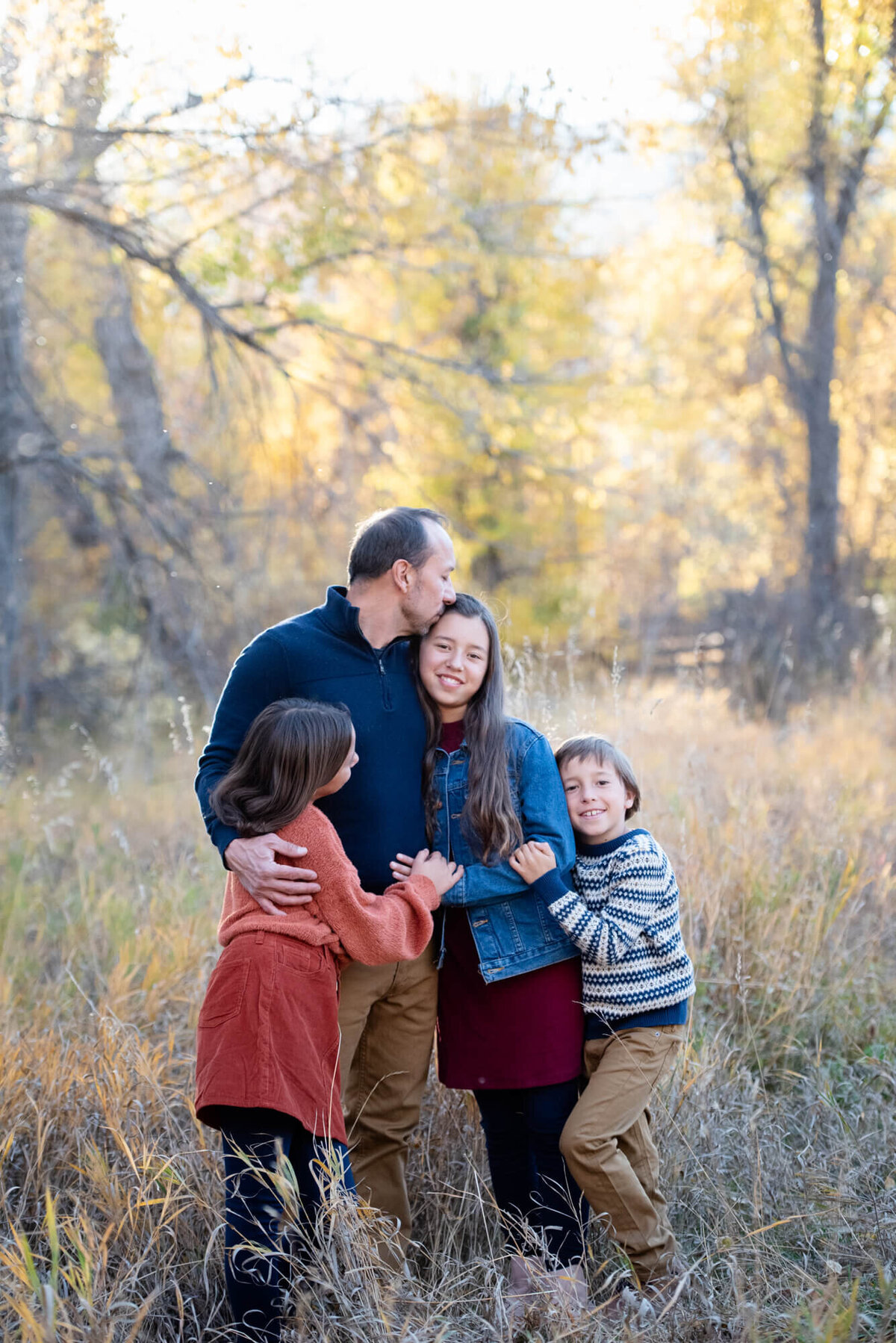 A father kisses his oldest daughter while being hugged by his younger son and daughter in a forest meadow for a Denver family photographer