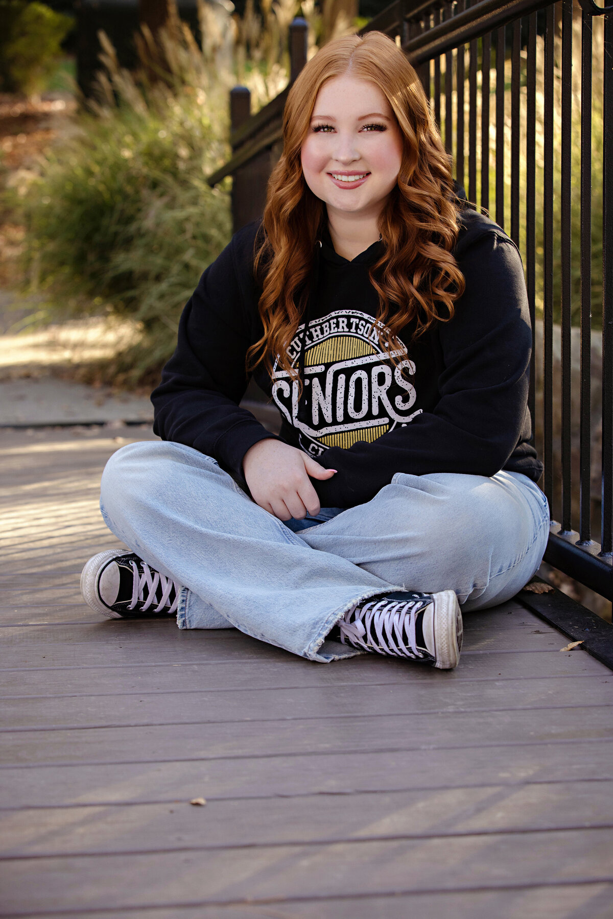 High school senior in black SENIORS hoodie sitting on wooden bridge over a lake, surrounded by trees