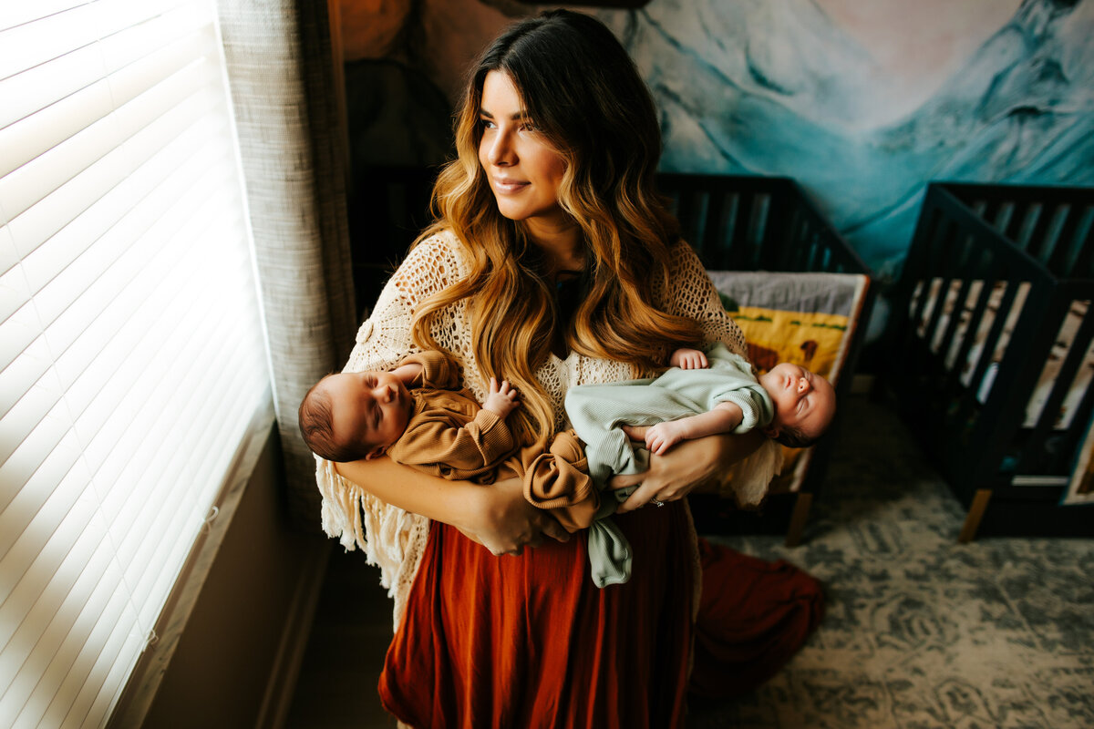 colorful newborn nursery photo session with a mom in a boho dress holding her newborn twin boys in front of a window