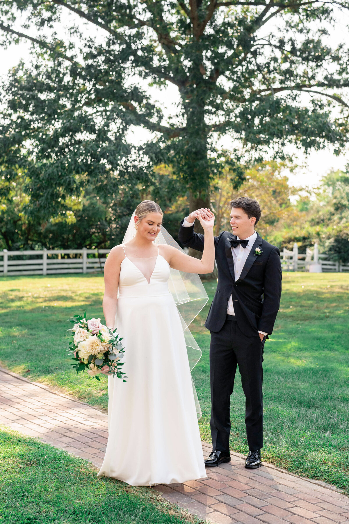 A bride in a white dress holds a bouquet of flowers and smiles as she dances with a groom in a black tuxedo. They are outdoors on a brick path, surrounded by greenery and trees.