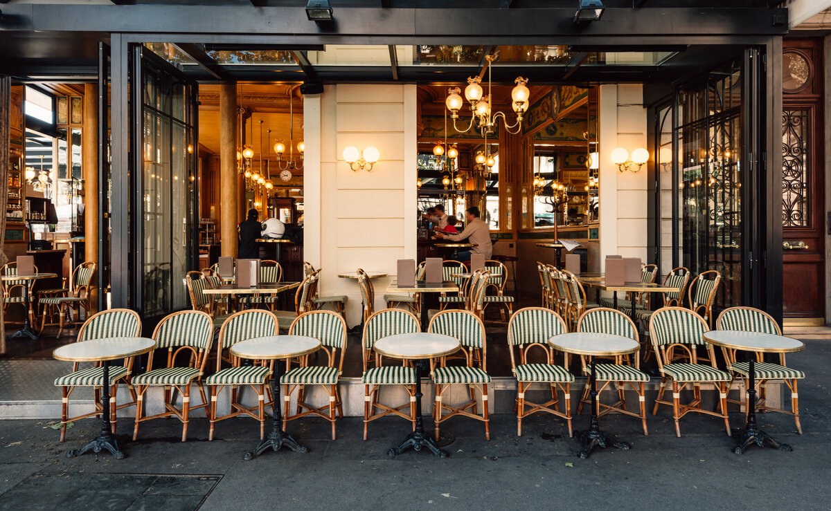 Front view of an open-air cafe with rows of round tables and striped chairs on the sidewalk. Warm lights illuminate the interior, where patrons can be seen enjoying the cozy ambiance. Large glass doors are open, welcoming guests inside.