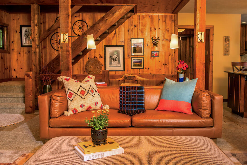 The cozy interior of a wood cabin with a prominent wooden staircase leading to the upper level. A comfortable brown leather sofa sits in the foreground with colorful pillows. The warm wood tones of the walls and staircase are complemented by soft lighting.