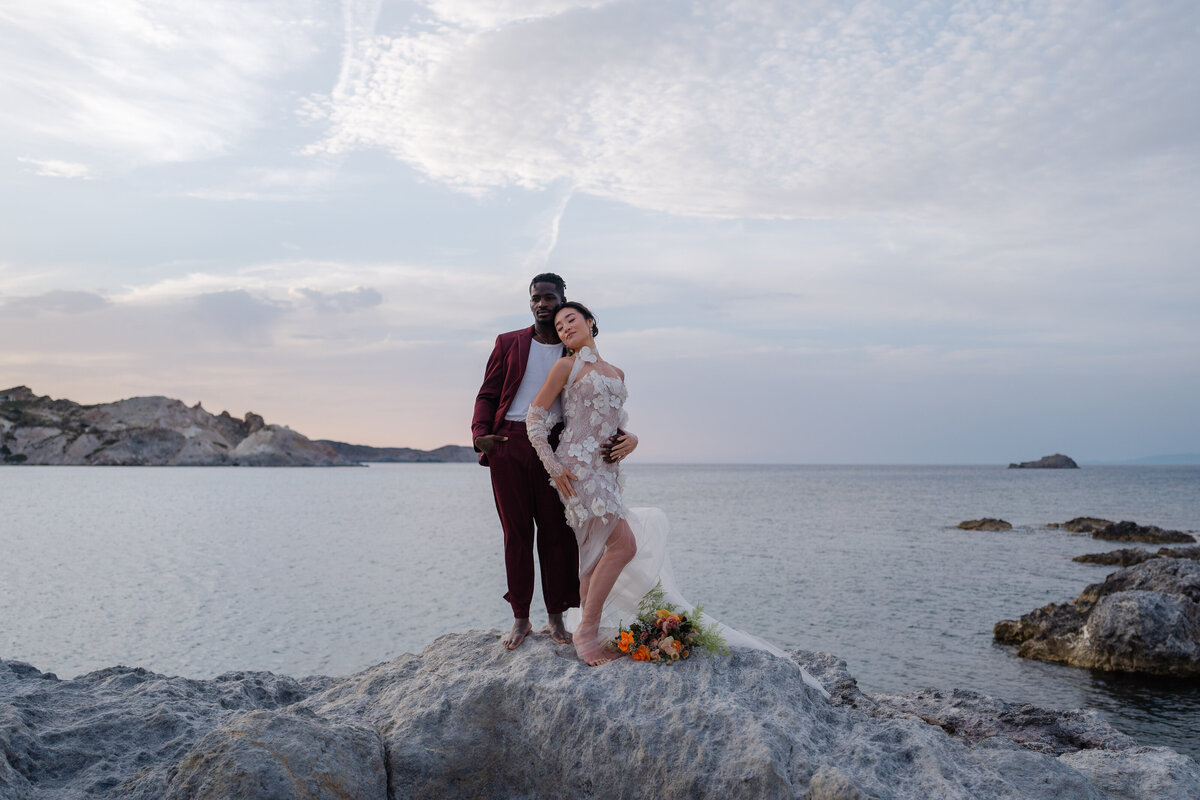 Multicultural couple barefoot on a rock in Milos, Greece, during a destination wedding photoshoot captured by Chi Ramos Photography.