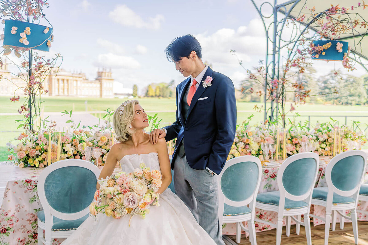 bride sitting in a blue chair at a pink and peach wedding dinner table looking up at her groom standing next to her with blenheim palace in the background