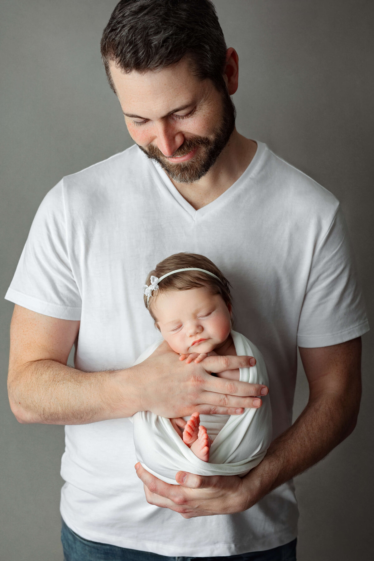 new father holding his newborn baby girl in a white shirt and wrap on a grey backdrop at a newborn photo session