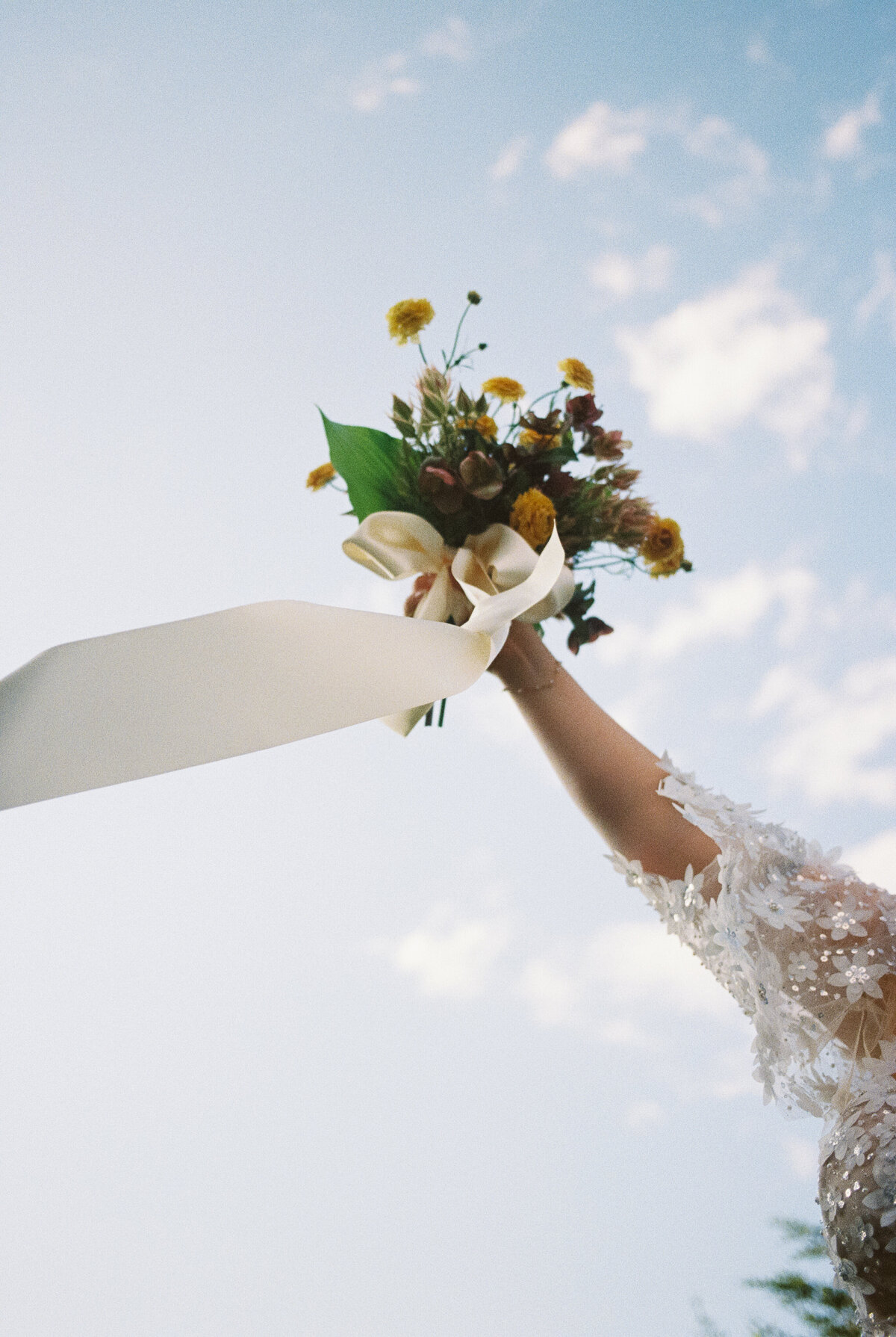 A bride holding out a yellow and green wedding bouquet into the air as the ribbon flies.