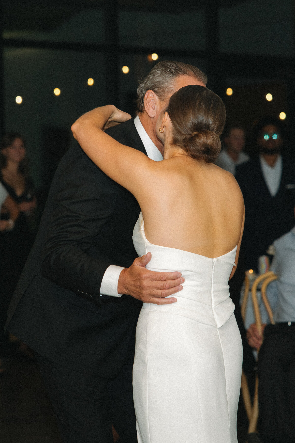 A bride in a white strapless, fitted wedding dress kisses her father on the cheek as she wraps her arms around her upper shoulders as they hug. Her father has a black suite with a white dress shirt underneath. People in formal attire stand and sit behind them watching them. 