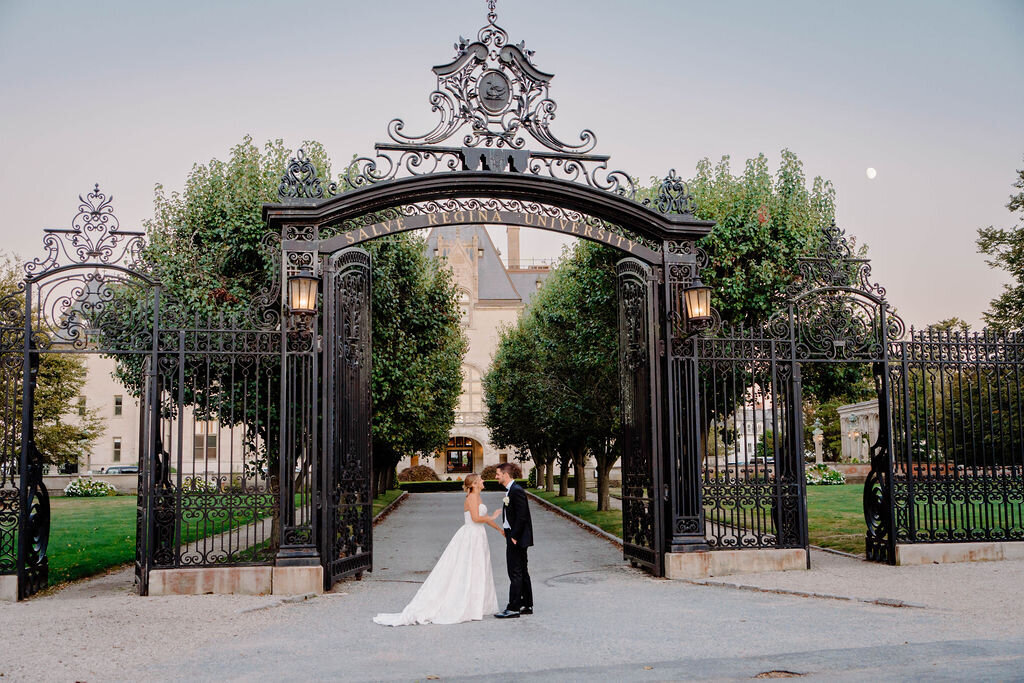 Ochre Court Newport | A bride and groom stand before an ornate, wrought-iron gate, embracing under a soft evening sky. Lush greenery and a grand building are visible behind them.