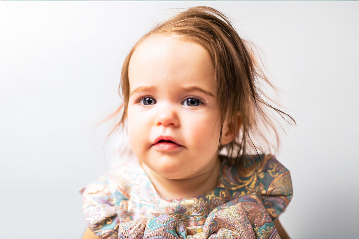 A one year-old blue-eyed baby girl looks directly at the camera as her beautiful we spoke children's outfit stands out against her creamy skin