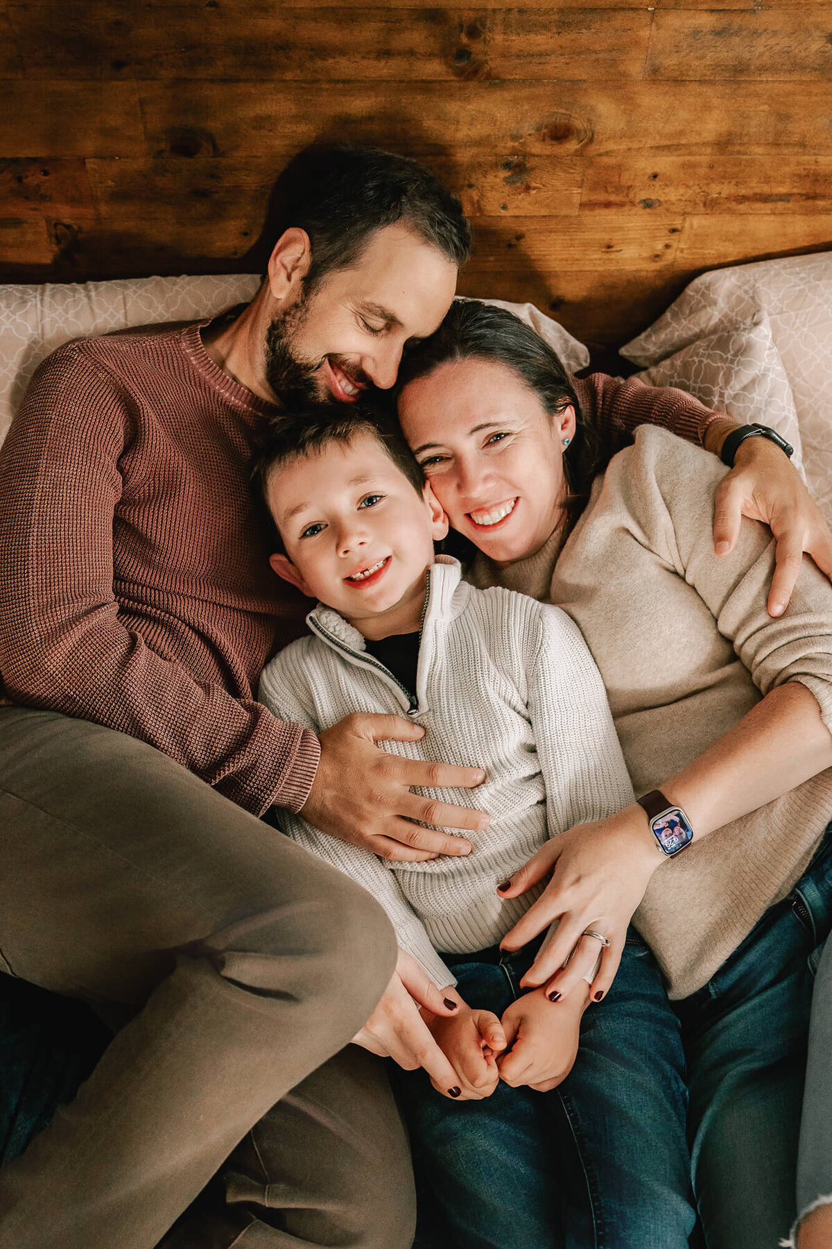 Bearded dad in a maroon sweater smiles and closes his eyes as he holds his wife and their 5 year old son close, all of them snuggled on the parents' bed.