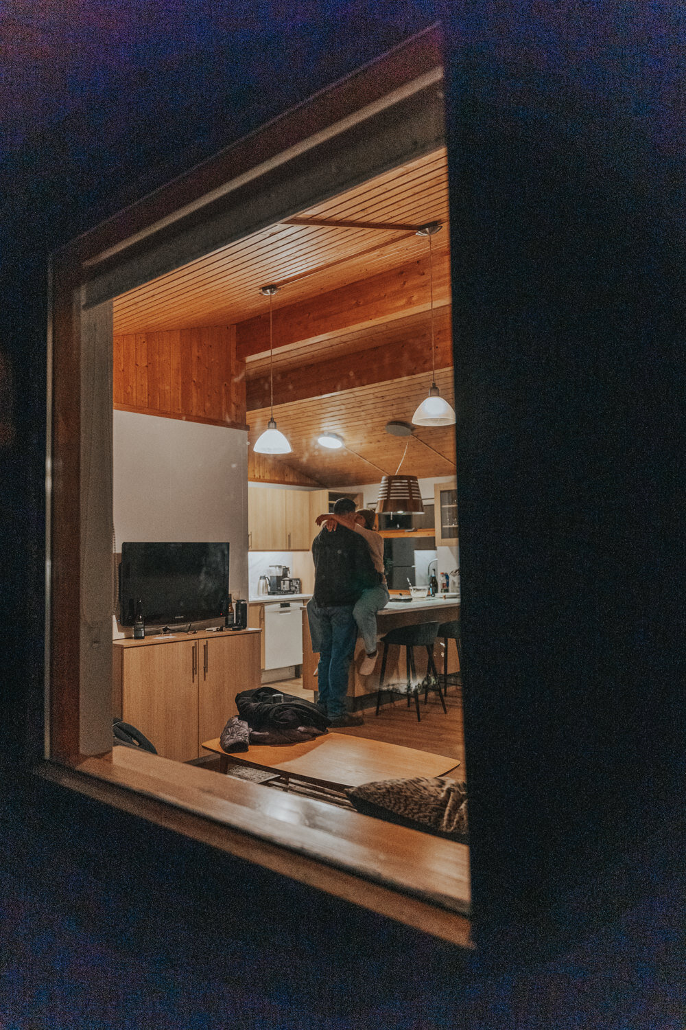 Woman sits on the bar of their kitchenette while holding her husband 