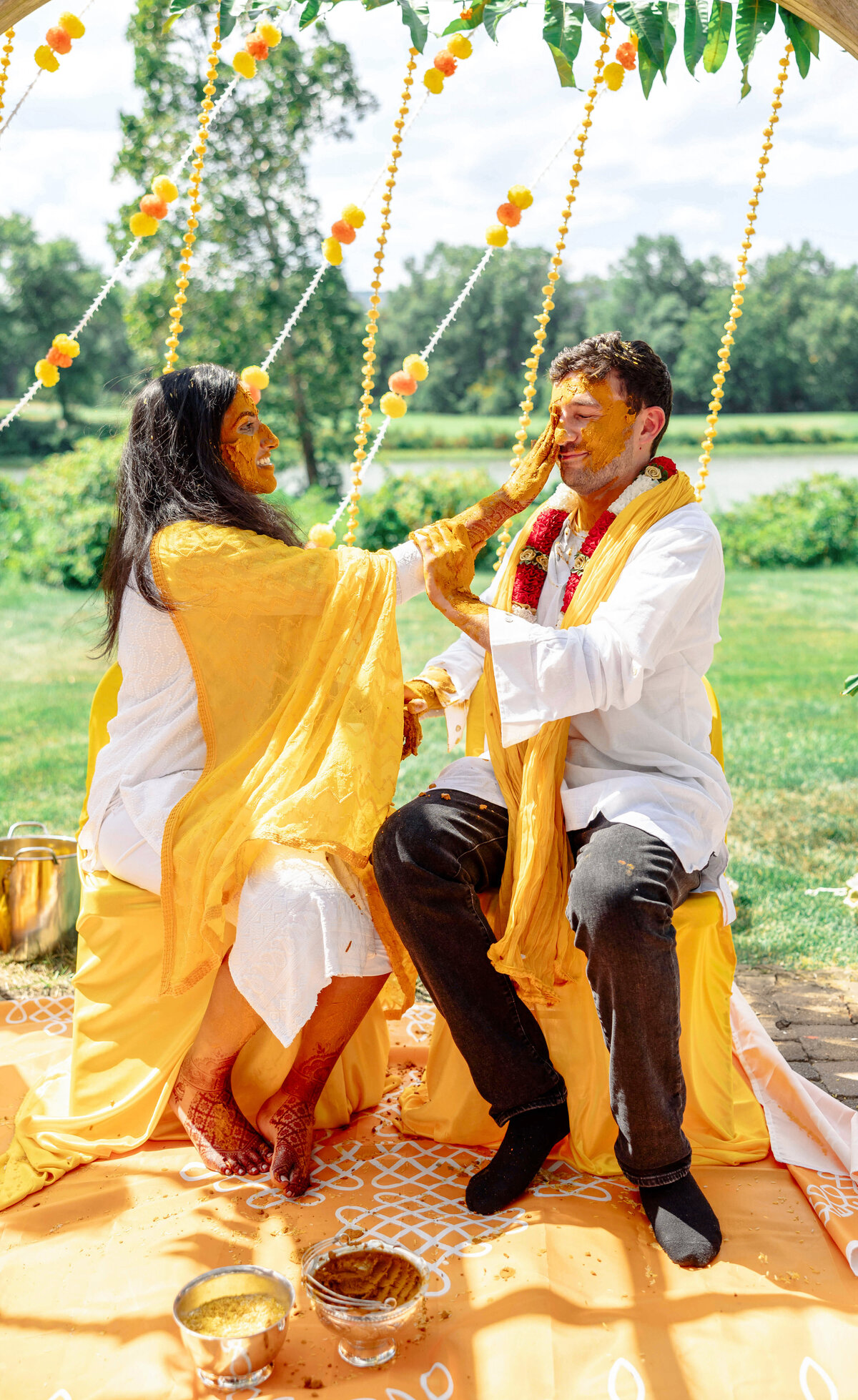 A couple participates in a traditional pre-wedding ceremony outdoors. They are playfully applying turmeric paste to each others faces. Both wear white outfits with yellow accents. Decorations with marigold flowers are in the background.