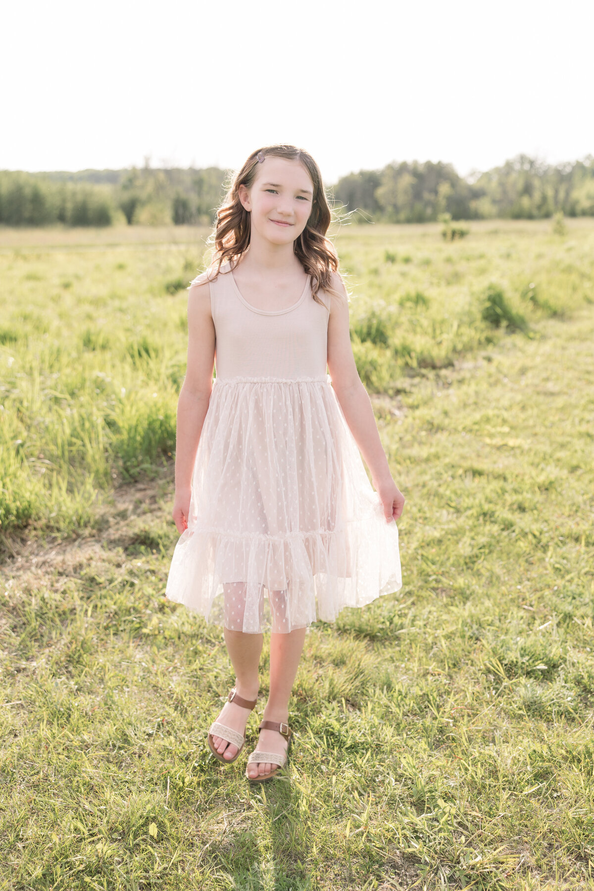 A little girl in a beige dress is walking toward the camera and smiling