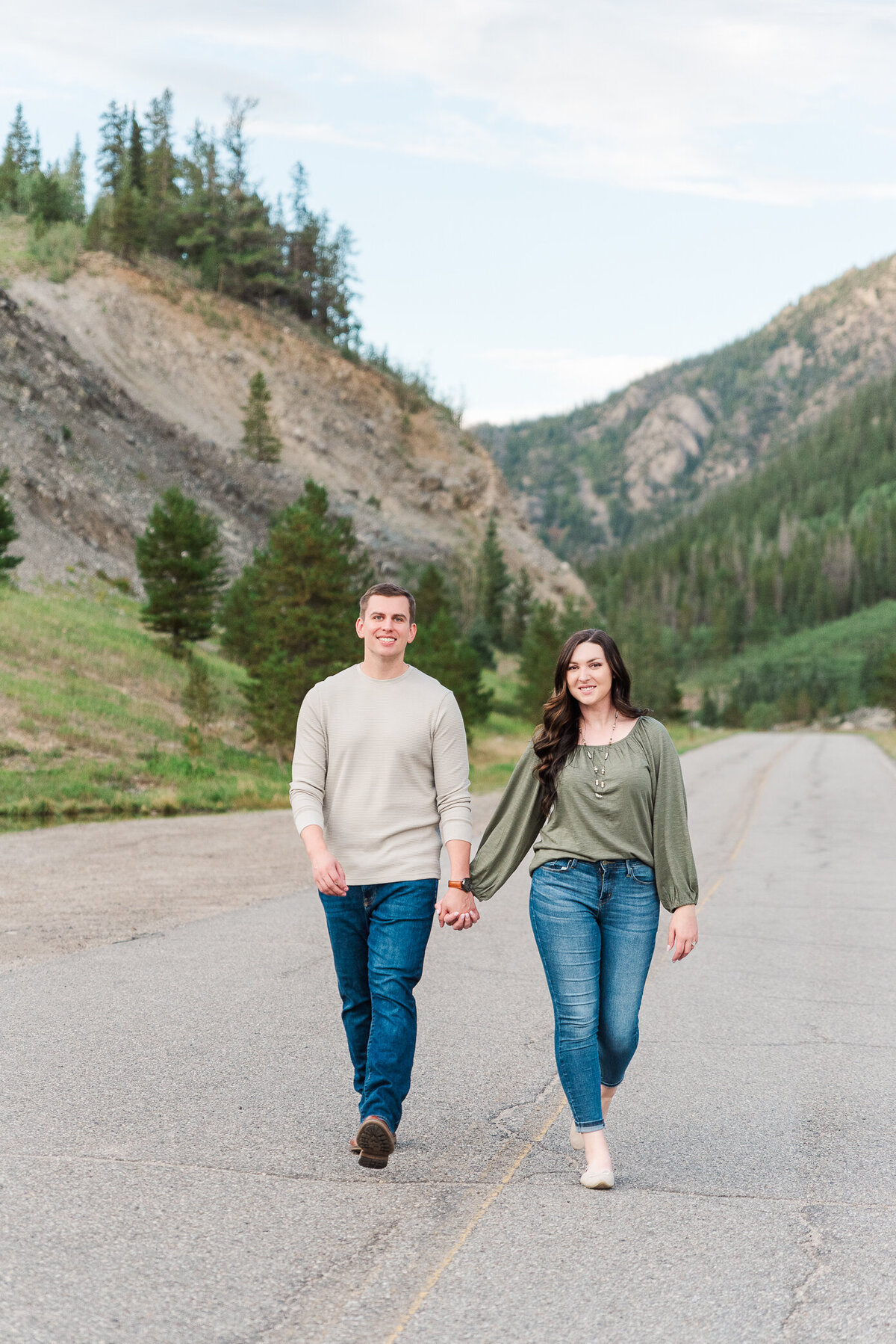 A woman in a green shirt and jeans walks hand in handwith her partner who is wearing a tan shirt and blue jeans walk along a mountain road together