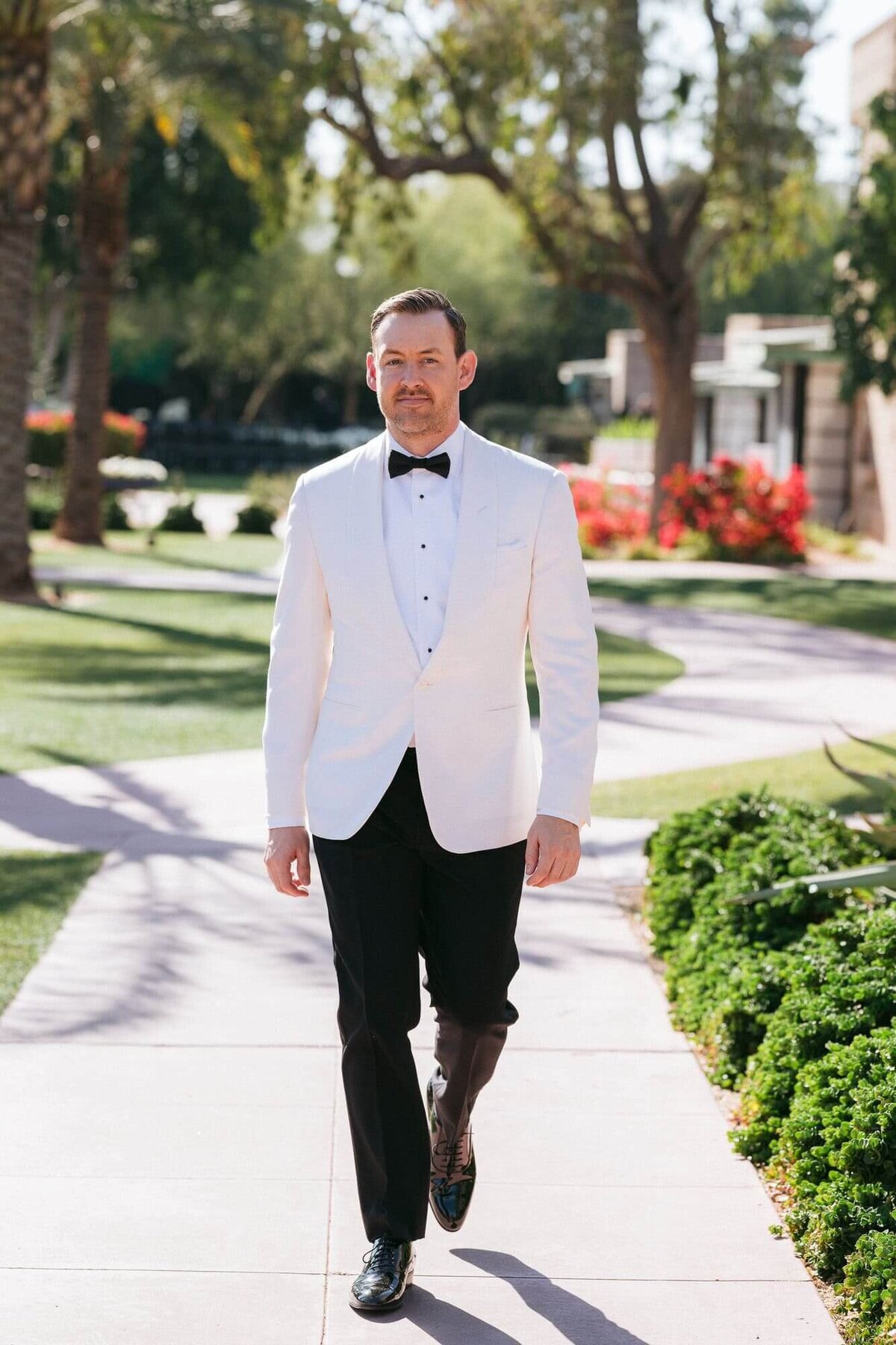Groom in a classic white tuxedo jacket and black bow tie walking the garden paths at Arizona Biltmore Scottsdale wedding venue.