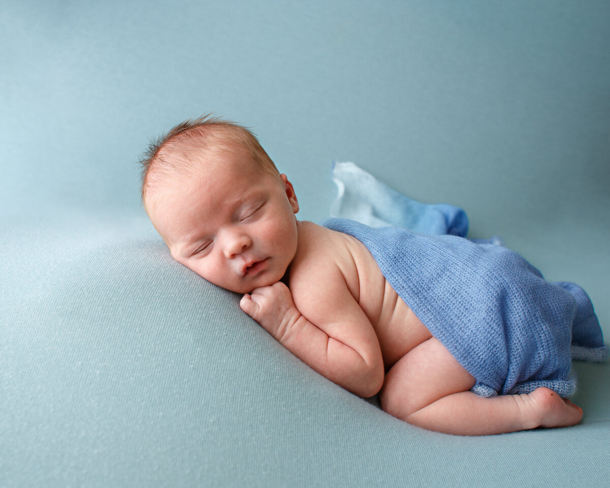 Portrait of an infant baby boy laying naked on his tummy with a blue blanket covering his bottom