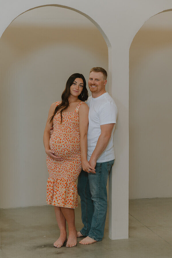 A man holds his pregnant wife from behind with one hand and her hand with the other. She's holding her pregnant belly as they both smile for the camera.