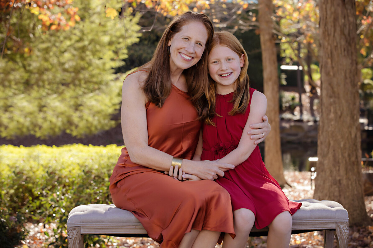 Sweet autumn portrait of mom and youngest daughter seated together on park bench surrounded by colorful leaves at Ballantyne Corporate Park