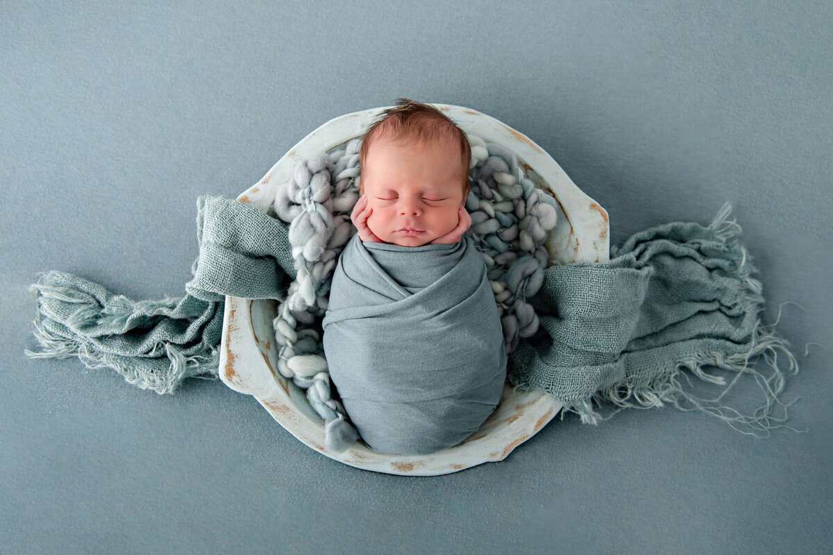 newborn boy wrapped in blue in a blue bowl on a blue background.