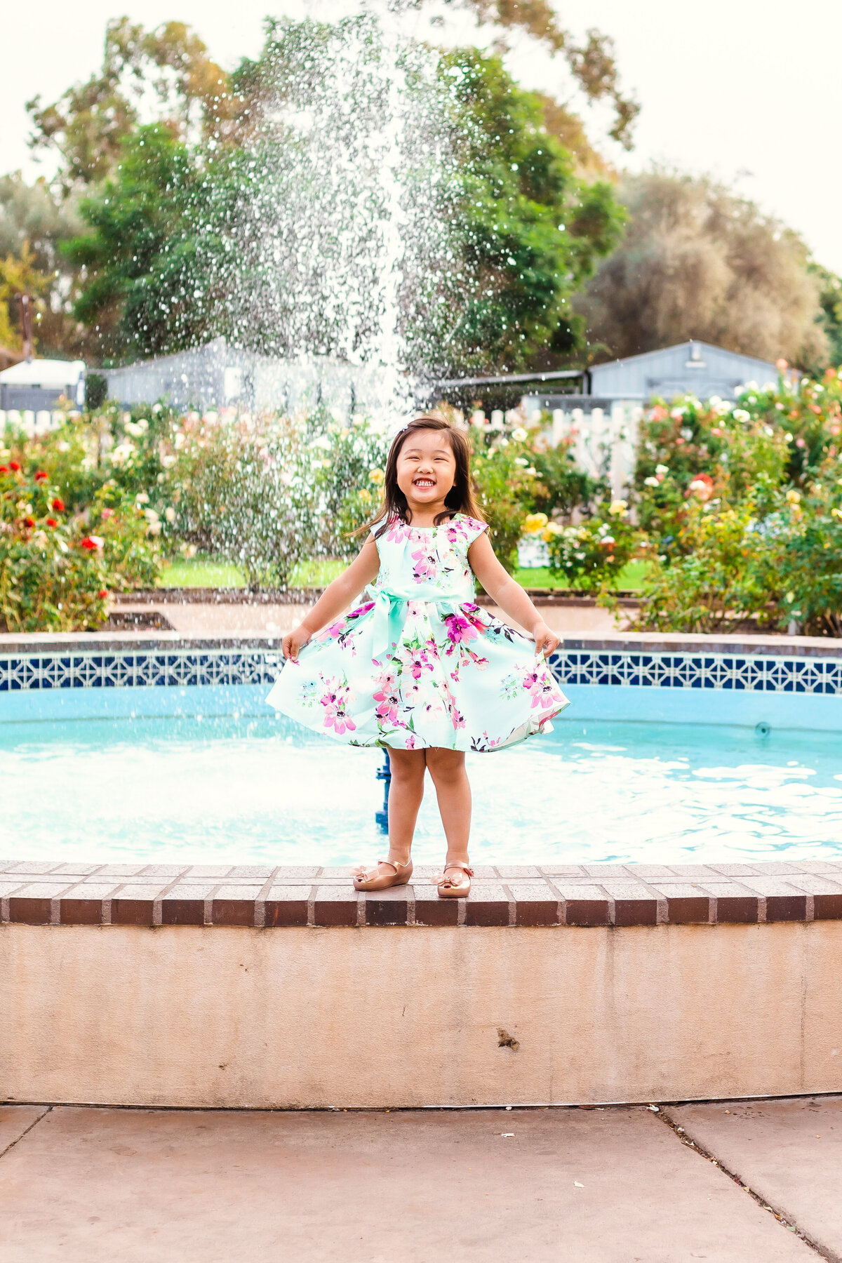 Family Photographer,  a little girl smiles and holds her dress before a fountain