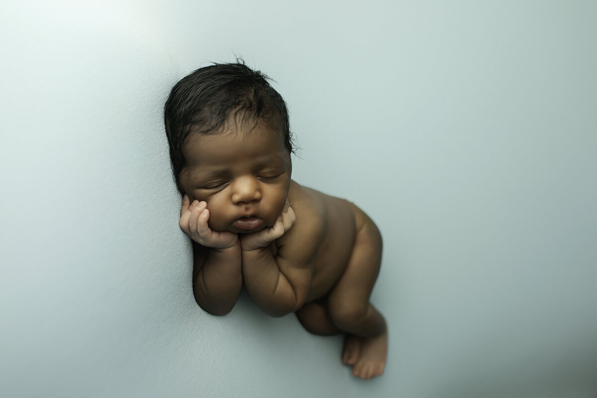 Newborn baby boy peacefully sleeping on a soft white backdrop, gently wrapped in a cozy grey fabric. His tiny hands peek out as he rests comfortably, with a serene expression and delicate features highlighted by soft, natural lighting.
