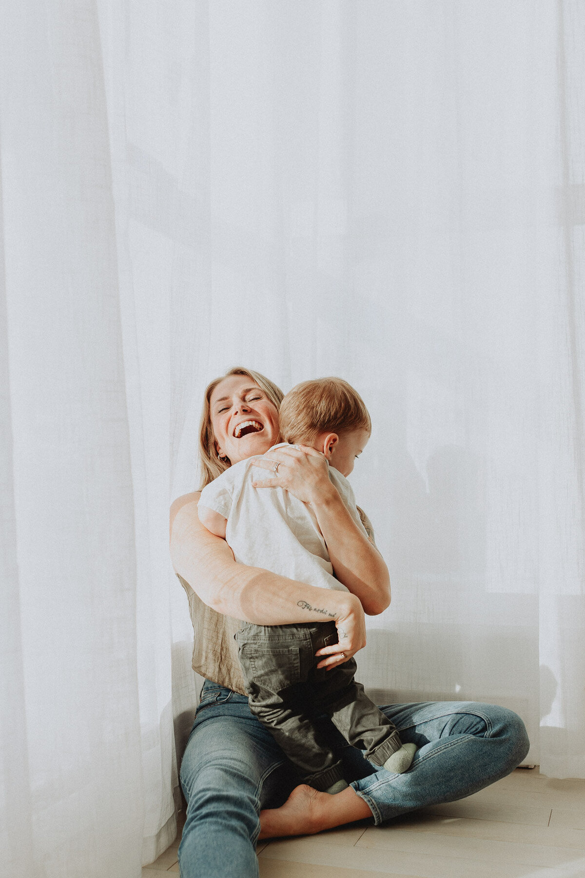 photo of a mom snuggling her toddler son sitting in front of bright white curtains with the sun shining through taken in Langley
