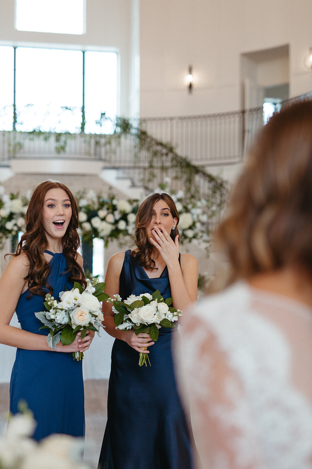 Two bridesmaids in blue satin gowns holding white bouquets gasp at bride in a banquet room filled with white florals.