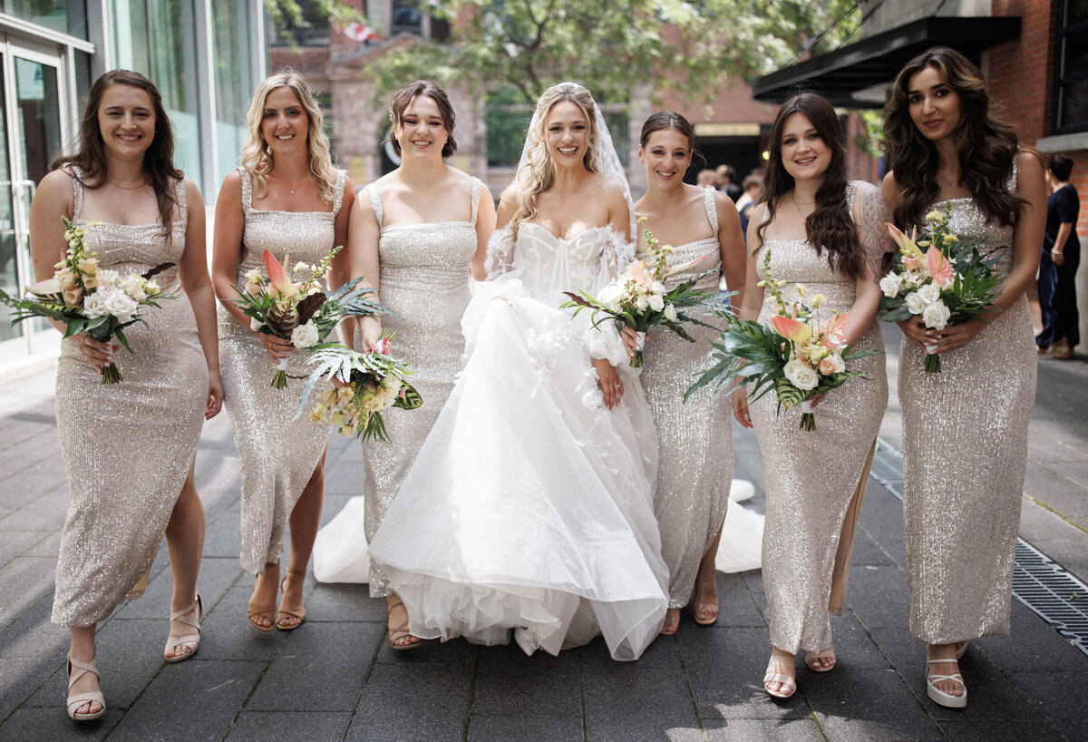 A group of bridesmaids holding exotic bouquets circle a bride in front of the Cathcart. 