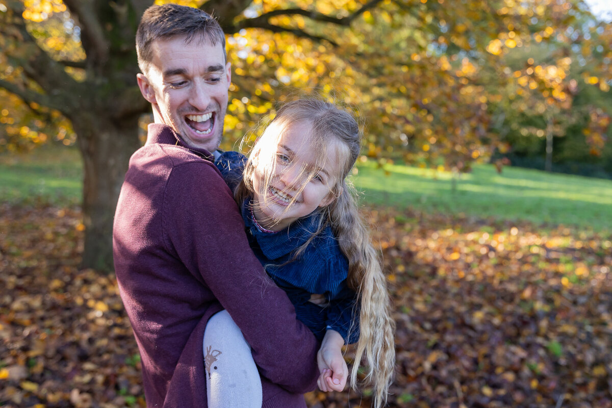 A man holding a young girl in his arms, both smiling, with a backdrop of trees showing autumn foliage.