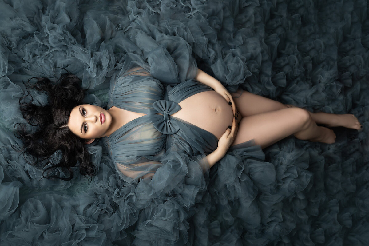 Woman surrounded by cascading teal tulle, lying gracefully with hands resting on her belly, dressed in a sheer gown with a bow detail. Maternity photography by Lindsay Walden Photography.