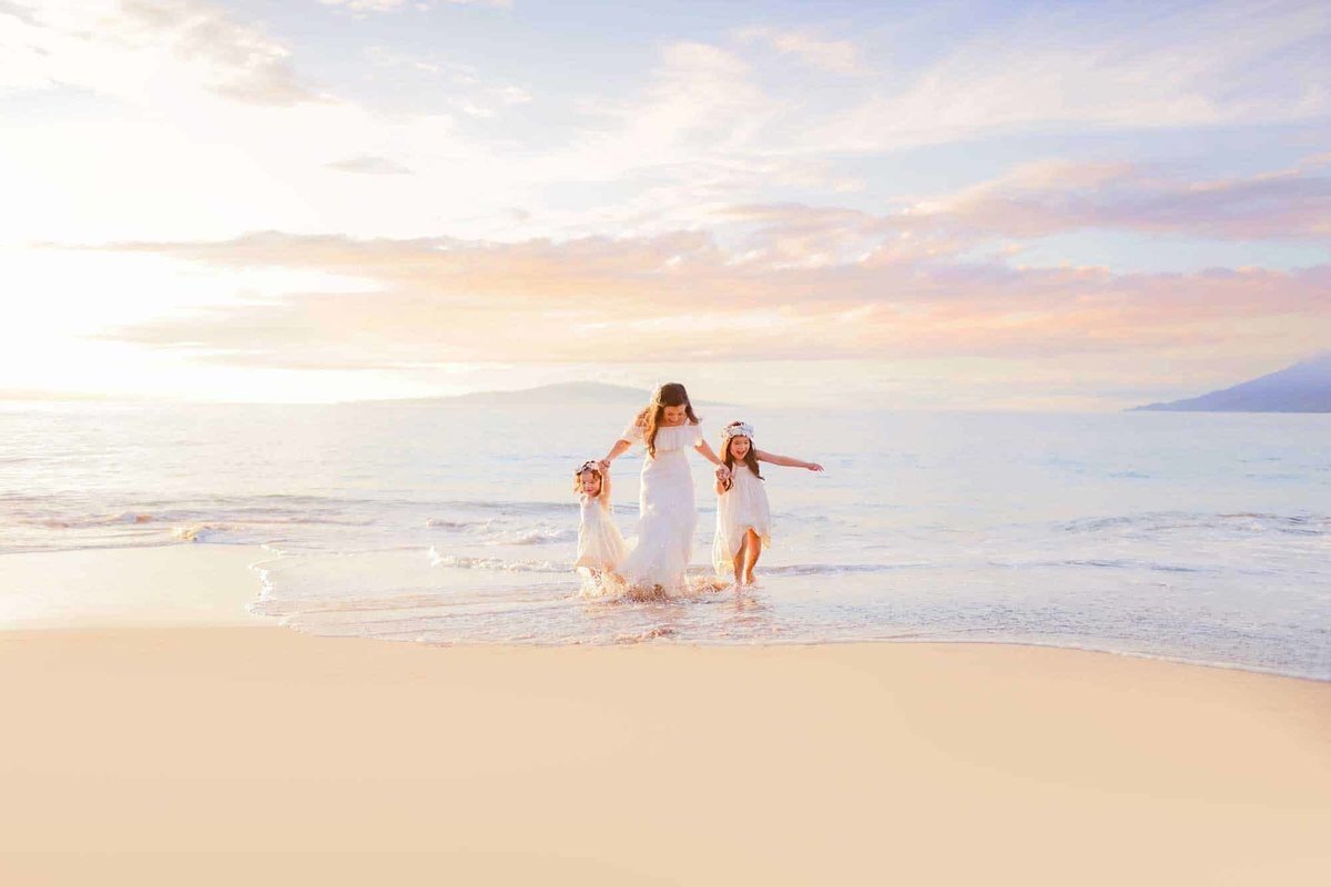 Mother and daughters wearing white dresses run hand-in-hand for their family beach photography session with Love + Water Photo