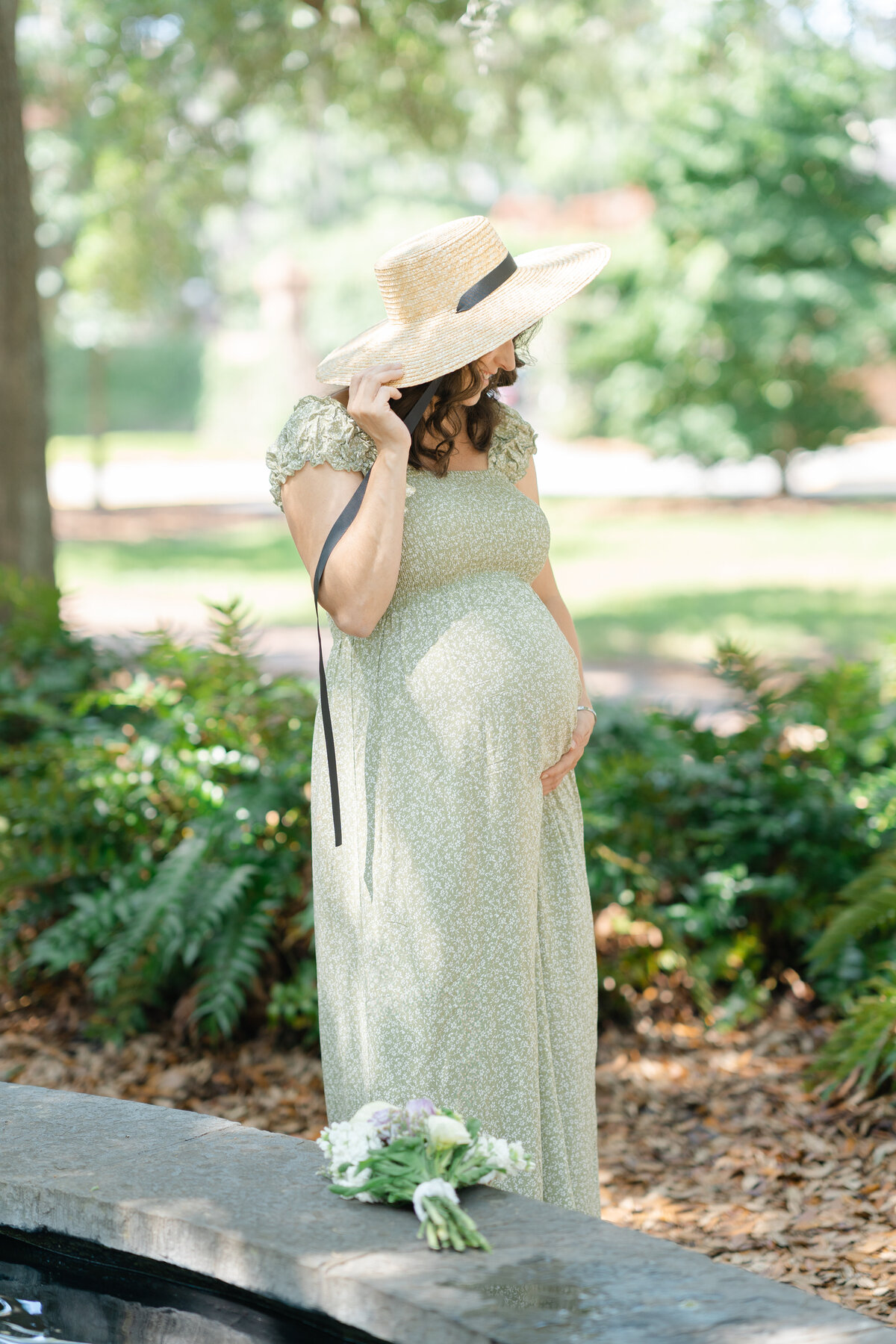 Pregnant mother in a flowy green dress and wearing a beautiful sun hat standing in Savannah square by Savannah family photographer Courtney Cronin