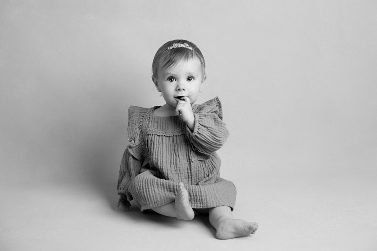 Six month old baby girl wearing a dress and looking sweetly at the camera with her finger in her mouth.