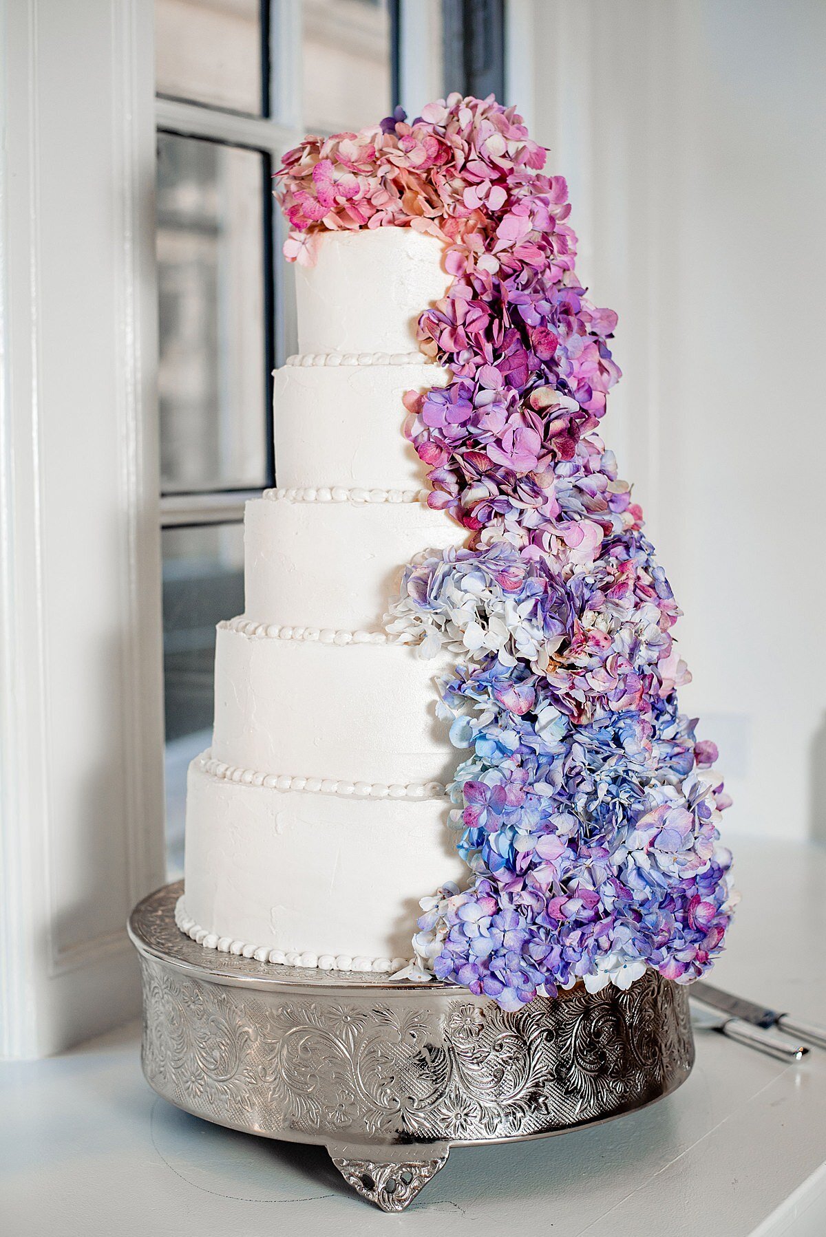 Five tier wedding cake with white pearls and a cascade of ombre hydrangea flowers ranging from pink hydrangea to purple hydrangea to blue hydrangea sitting on a silver cake stand at Noelle Hotel Nashville